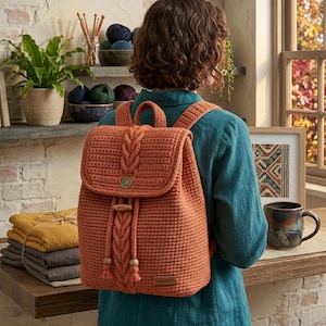 May include: A burnt orange crochet backpack featuring a braided cable knit pattern on the front flap and center panel. The bag includes a wooden toggle closure, two shoulder straps, and a small rectangular tag labeled handmade on the bottom corner.