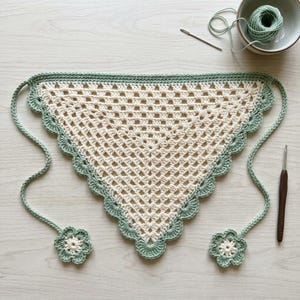 May include: A triangular crochet bandana featuring a cream-colored granny square pattern with a sage green scalloped border. The piece includes two long ties, each finished with a small flower motif at the end, crafted from matching yarn.