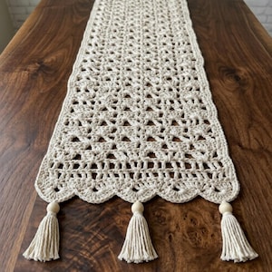 May include: A cream-colored rectangular table runner featuring an intricate openwork crochet pattern with a scalloped edge. The runner is finished with three matching cream-colored tassels, each topped with a smooth, round, light-colored wooden bead.