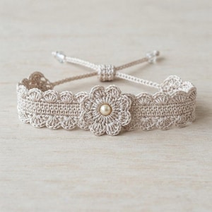 May include: A beige crochet bracelet featuring a central flower motif with a single cream-colored pearl at its core. The band has a scalloped edge design and includes adjustable thin cord ties with small clear beads at the ends for a secure fit.