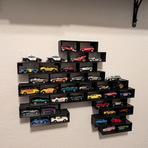 May include: A black wall-mounted display shelf with an irregular, stepped geometric shape, featuring multiple rectangular compartments. The shelf holds a collection of various miniature toy cars in diverse colors, styles, and designs.