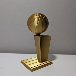 May include: A gold-colored trophy featuring a basketball perched atop a tapered, angular pedestal base. The entire structure is monochromatic with a smooth, metallic finish, resting on a rectangular platform with visible horizontal lines.