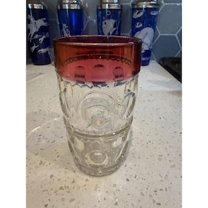 May include: A clear glass tumbler featuring a deep red band around the top rim and a textured pattern of circular indentations along the sides. The glass has a thick base and a cylindrical shape, showcasing a distinct contrast between the red rim and the clear body.