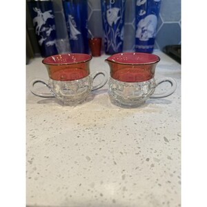 May include: Two clear glass creamers with a textured, faceted base and a smooth, deep red rim. Each creamer features a single curved handle extending from the side. The glass has a reflective quality, showing a distinct contrast between the clear bottom and the colored top section.