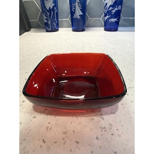 May include: A square-shaped red glass bowl with rounded corners and a smooth, translucent finish. The interior base of the bowl features a subtle, radiating sunburst pattern pressed into the glass, creating a clear focal point at the center.