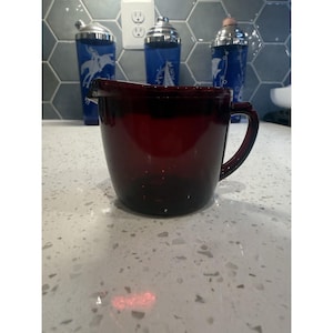 May include: A deep red glass pitcher with a rounded body, a short spout, and a sturdy side handle. The pitcher features a smooth, translucent finish and is positioned in the foreground, showcasing its dark crimson hue and simple, functional design.