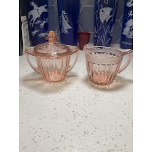 May include: A pink depression glass sugar bowl with a matching lid and a creamer pitcher. Both pieces feature a vertical ribbed pattern on the body and a delicate embossed floral design around the upper rim and the lid handle.