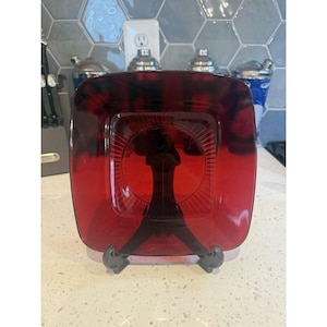 May include: A square, translucent ruby red glass plate featuring a central circular indentation with radiating linear patterns. The plate is displayed upright on a black plastic stand, showcasing its deep red color and smooth, reflective surface.