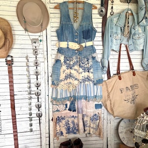 May include: A sleeveless denim vest with a button-up front and a cream-colored belt, layered over a multi-textured skirt featuring blue and white toile patterns, striped fabric, and lace trim. The garment is displayed on a wooden hanger.