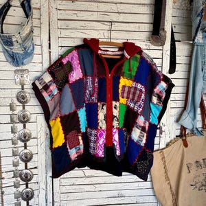 May include: A zip-up hooded poncho featuring a patchwork design made of various fabric squares in colors including red, blue, yellow, green, and black. The garment has a red hood with drawstrings and a central red zipper closure.