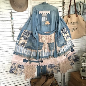 May include: A light blue denim jacket with patchwork details, including floral and lace accents. The jacket features a unique design with a flowing, layered skirt and a decorative back panel. The overall style is bohemian and vintage-inspired.