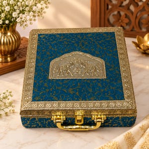May include: A rectangular blue box with a gold floral pattern and a metallic gold border. The lid features a central arched plaque depicting two embossed elephants. The box includes a gold metal handle and a central latch closure mechanism.