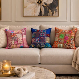 May include: Three square decorative throw pillows featuring intricate patchwork designs with embroidery, sequins, and beadwork. The pillows are colored in vibrant shades of pink, deep blue, and bright orange, each displaying a unique grid pattern.