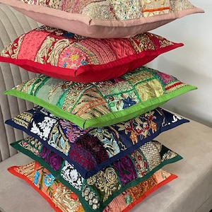 Könnte beinhalten: Ein Stapel aus sechs quadratischen Zierkissen mit Patchwork-Designs, aufwendigen Stickereien, metallischen Garnakzenten und verschiedenen Stofftexturen in Beige, Rot, Grün, Blau, Lila und Orange, jeweils mit einfarbiger Paspelierung.