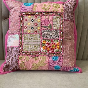 May include: A square throw pillow featuring a patchwork design in shades of pink, gold, and teal. The surface is composed of various fabric panels with intricate embroidery, floral patterns, small mirrors, and decorative stitching throughout.