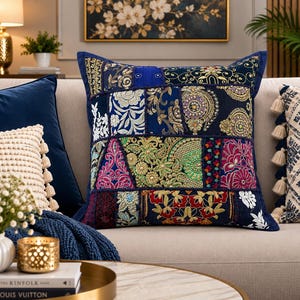 May include: A square throw pillow featuring a patchwork design of dark blue fabric panels. Each panel is decorated with intricate embroidery in gold, white, pink, and green thread, incorporating floral motifs, geometric patterns, and small reflective accents.