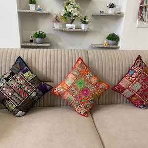 以下が含まれることがあります： 刺繍、スパンコール、ビーズ細工が施された複雑なパッチワークデザインの正方形のクッション3つ。左のクッションは主に黒、中央はオレンジ、右は濃い赤で、ベージュのソファの上に置かれています。