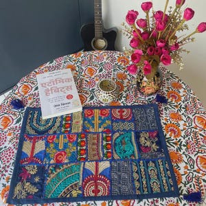 May include: A rectangular patchwork table runner featuring various embroidered fabric squares in shades of blue, gold, red, and green, placed on a round table with a white floral print cloth, a book titled Atomic Habits, and a vase of pink flowers.