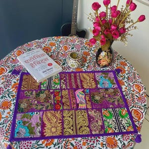 May include: A rectangular fabric table runner featuring a patchwork of purple, gold, and green embroidered panels with floral and bird motifs, accented by purple tassels at the corners, resting on a white tablecloth with a vibrant orange and green floral print.