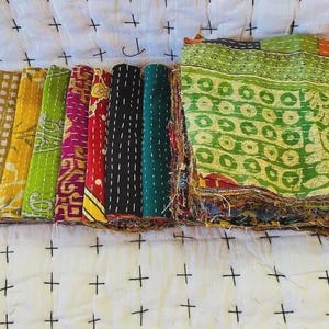 Peut inclure: Une collection de carrés de tissu pliés présentant divers motifs et couleurs, notamment le vert, le jaune, le rouge et le noir. Chaque pièce est cousue avec un fil blanc, créant un aspect texturé. La pile affiche plusieurs couches de textiles à motifs avec des bords effilochés, mettant en valeur une variété de designs et de teintes dans un agencement compact et organisé.