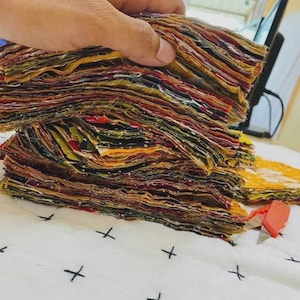 Op de afbeelding: Een grote stapel vierkante stofresten met een verscheidenheid aan kleuren, waaronder geel, rood, groen en zwart. De stukken stof zijn netjes op elkaar gestapeld en tonen rauwe randen en diverse patronen. Een klein oranje hobbymesje ligt ernaast.