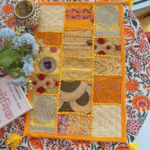 May include: A rectangular wall hanging featuring a patchwork design of yellow, gold, and cream fabric squares. The piece includes intricate embroidery, beadwork, and floral patterns, finished with a bright yellow border and yellow corner tassels.