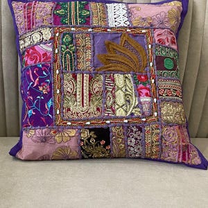 Könnte beinhalten: Ein quadratisches Zierkissen mit einem Patchwork-Design aus verschiedenen Stoffresten in den Farben Violett, Rosa, Gold und Grün. Die Oberfläche zeigt aufwendige Stickereien, Details aus Metallic-Garn, florale Muster und ein zentrales Lotus-Motiv.