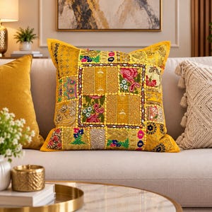 Puede incluir: Un cojín decorativo cuadrado con un vibrante diseño de patchwork amarillo. La superficie está compuesta por varios paneles de tela bordados con intrincados patrones florales, detalles en hilo dorado metálico y pequeños espejos.