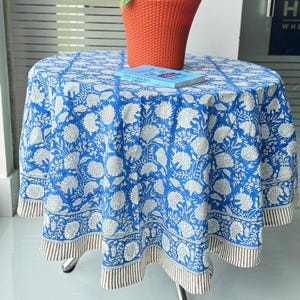Puede incluir: Una mesa redonda cubierta con un mantel de estampado floral azul y blanco con un borde a rayas. Sobre la mesa hay una maceta de plástico naranja con una planta verde y un libro titulado Think & Grow Rich con una cubierta azul.