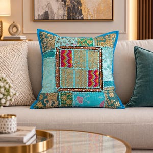 Peut inclure: Un coussin carré présentant un motif patchwork dans des tons turquoise, bleu sarcelle et doré. Le carré central est encadré par des bordures brodées avec des motifs en zigzag roses et dorés, entouré de panneaux de broderies florales et dorées.