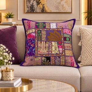 Puede incluir: Un cojín cuadrado con un diseño de patchwork vibrante que presenta varios paneles de tela en tonos morados, rosas y dorados. El cuadrado central muestra bordados intrincados y detalles en hilo dorado, rodeado por un borde morado con pequeñas cuentas blancas.