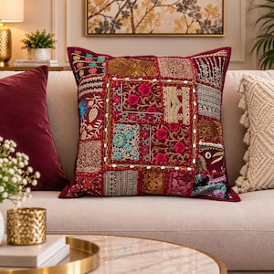 May include: A square throw pillow featuring a patchwork design of deep red fabric panels. The surface is decorated with intricate embroidery, metallic gold thread accents, floral patterns, and small white beads arranged in a grid-like layout.