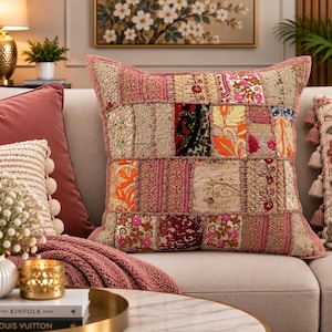 May include: A square throw pillow featuring a patchwork design of various embroidered fabrics in shades of pink, gold, orange, and red. The surface displays intricate floral patterns, metallic thread accents, and beadwork, finished with a pink border.