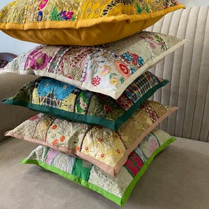 Pode incluir: Uma pilha de cinco almofadas quadradas com designs de patchwork, apresentando bordados intrincados, missangas e detalhes em fio metálico. As almofadas exibem uma variedade de cores, incluindo amarelo, branco, verde e bege com padrões florais.