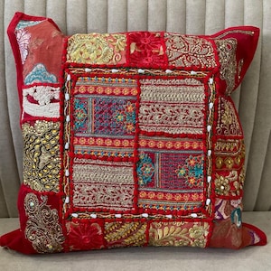 Könnte beinhalten: Ein quadratisches Zierkissen mit einem Patchwork-Design aus roten Stoffbahnen. Die Oberfläche ist mit aufwendigen Goldstickereien, bunten Blumenmustern, metallischen Fäden und kleinen weißen Perlen entlang des zentralen quadratischen Randes verziert.