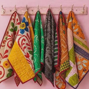 May include: Six folded quilted fabric pieces hang from a wooden rack. Each piece features distinct patterns and colors, including floral motifs, geometric shapes, and stripes in shades of yellow, green, red, black, and orange with visible stitching.