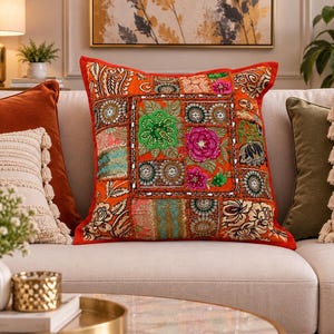 Peut inclure: Un coussin carré avec une base orange vif et un motif patchwork. La surface est ornée de broderies complexes, de motifs floraux verts et roses, ainsi que de motifs circulaires agrémentés de petites paillettes réfléchissantes.