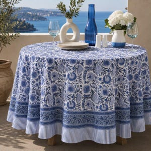 May include: A round tablecloth featuring a white base with a blue floral pattern, a decorative border, and a striped hem. The fabric drapes over a circular table, which is set with a stack of plates, a blue glass bottle, two wine glasses, and a vase.