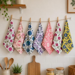 May include: Seven rectangular kitchen towels hanging from a horizontal twine string with wooden clothespins. Each towel features a distinct floral pattern in various color combinations including pink, blue, purple, and yellow on white backgrounds.