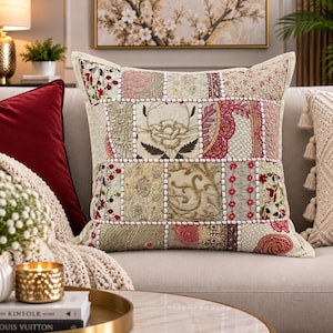 Pode incluir: Uma almofada decorativa quadrada com um design de patchwork de vários tecidos bordados em tons de creme, bege, rosa e vermelho escuro. Os painéis são unidos por costuras brancas, exibindo padrões florais e desenhos intrincados.