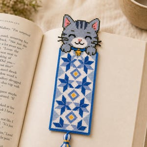 May include: A rectangular cross-stitch bookmark featuring a gray cat head at the top with a blue collar and yellow bell. The body of the bookmark displays a repeating geometric star pattern in shades of blue, white, and yellow, finished with a tassel.