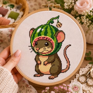 Op de afbeelding: Een kruissteek borduurproject in een ronde houten borduurring met een bruine muis die een groene watermeloen-kap draagt met een klein belletje. Het ontwerp is geborduurd op witte stof met een lichtbruine buik en roze oren.