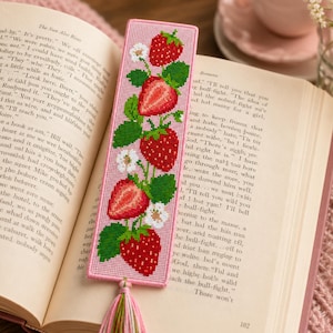 May include: A rectangular cross-stitch bookmark with a light pink border and background, featuring a vertical pattern of red strawberries, green leaves, and small white flowers. A multi-coloured tassel in shades of pink, green, and white is attached.