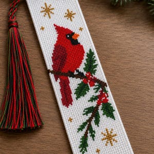 May include: A white cross-stitch bookmark featuring a red cardinal bird perched on a green holly branch with red berries. The design includes small gold star accents and is finished with a long, multi-coloured tassel in red, green, and black.