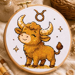 May include: A cross-stitch embroidery of the Taurus zodiac sign, depicting a smiling bull with large horns in gold and brown hues. The design is set against a white backdrop and framed in a wooden hoop. Decorative stars surround the bull.