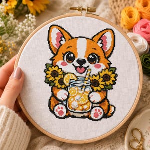 Può includere: Un progetto di ricamo a punto croce in un telaio di legno rotondo raffigurante un corgi dei cartoni animati con pelo arancione e bianco. Il corgi tiene in mano un barattolo di vetro con una bevanda al limone gialla, circondato da girasoli gialli.
