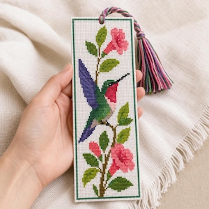 Peut inclure: Un marque-page rectangulaire en point de croix sur tissu blanc représentant un colibri à la gorge rouge, au corps vert et aux ailes violettes, perché sur une branche avec des fleurs roses en trompette et des feuilles vertes, avec un pompon multicolore.