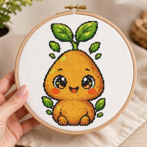 May include: A cross-stitch embroidery project in a round wooden hoop featuring a pixel-art style orange pear-shaped character with large black eyes, a small smile, pink cheeks, and green leaves sprouting from the top and sides on white fabric.