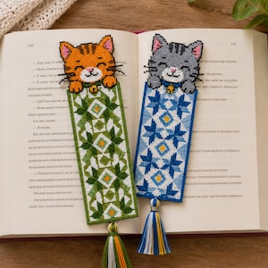 Puede incluir: Dos marcapáginas rectangulares de punto de cruz con cabezas de gato en la parte superior. Uno tiene un gato naranja con un patrón geométrico verde y una borla verde, blanca y naranja. El otro tiene un gato gris con un patrón geométrico azul y una borla azul, blanca y amarilla.