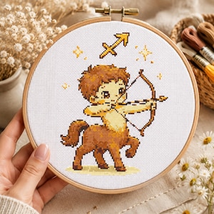 May include: A cross-stitch embroidery of the Sagittarius zodiac sign, featuring a cartoon centaur with a bow and arrow. The design is in shades of brown and yellow, set against a white background within a wooden hoop. Decorative elements include stars and the Sagittarius symbol.